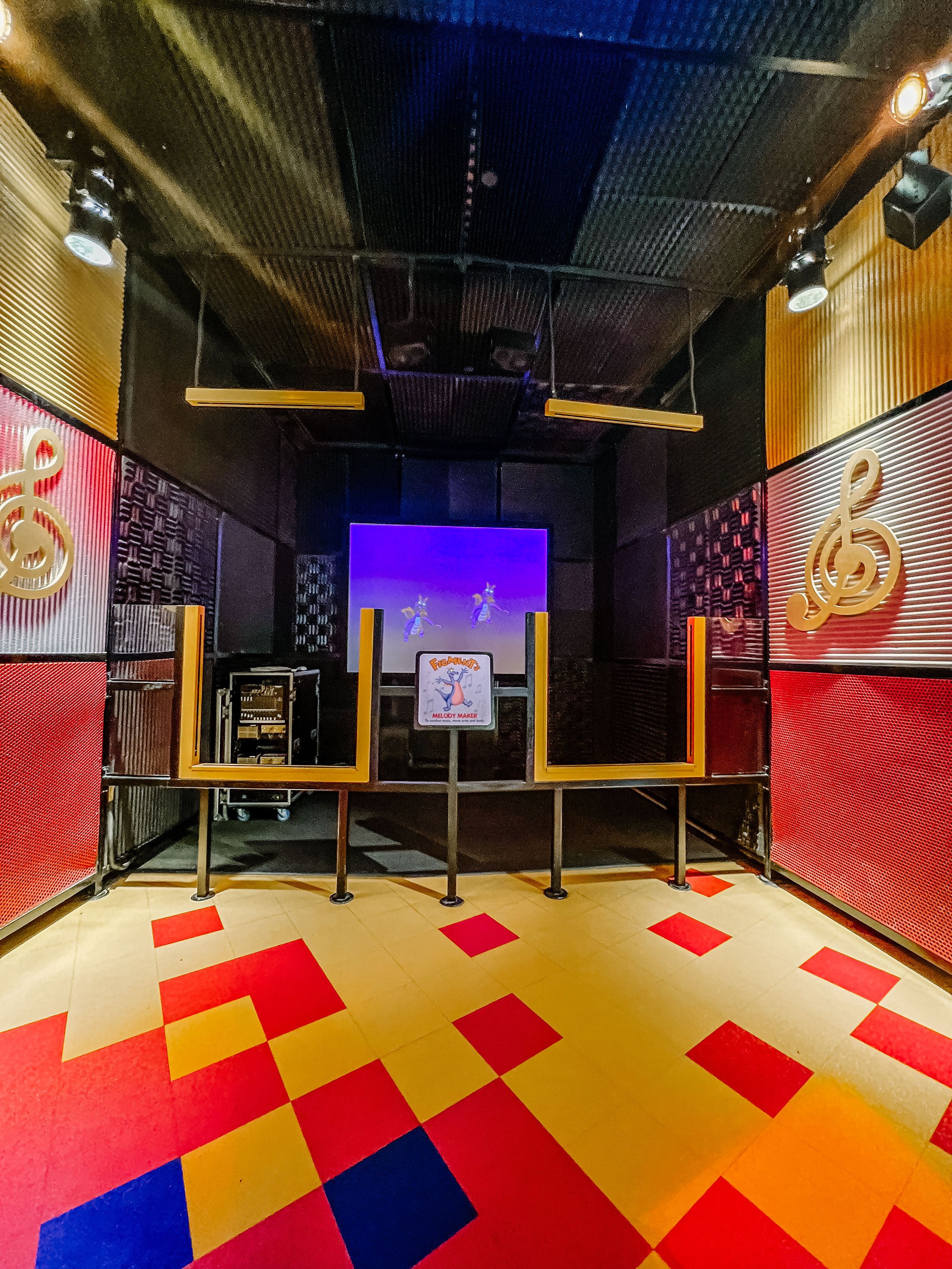 A colorful karaoke room with red and yellow checkered floor, LED panels on walls, and stage setup with screen and barriers.