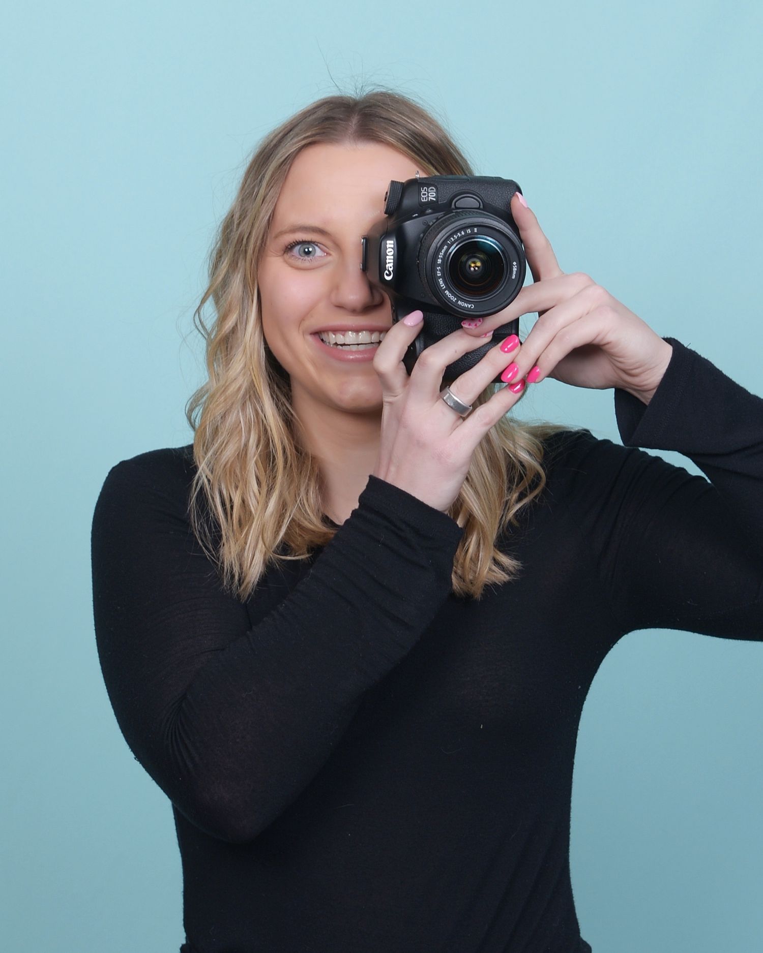 A photographer in a black sweater holds up a DSLR camera while smiling against a blue background.