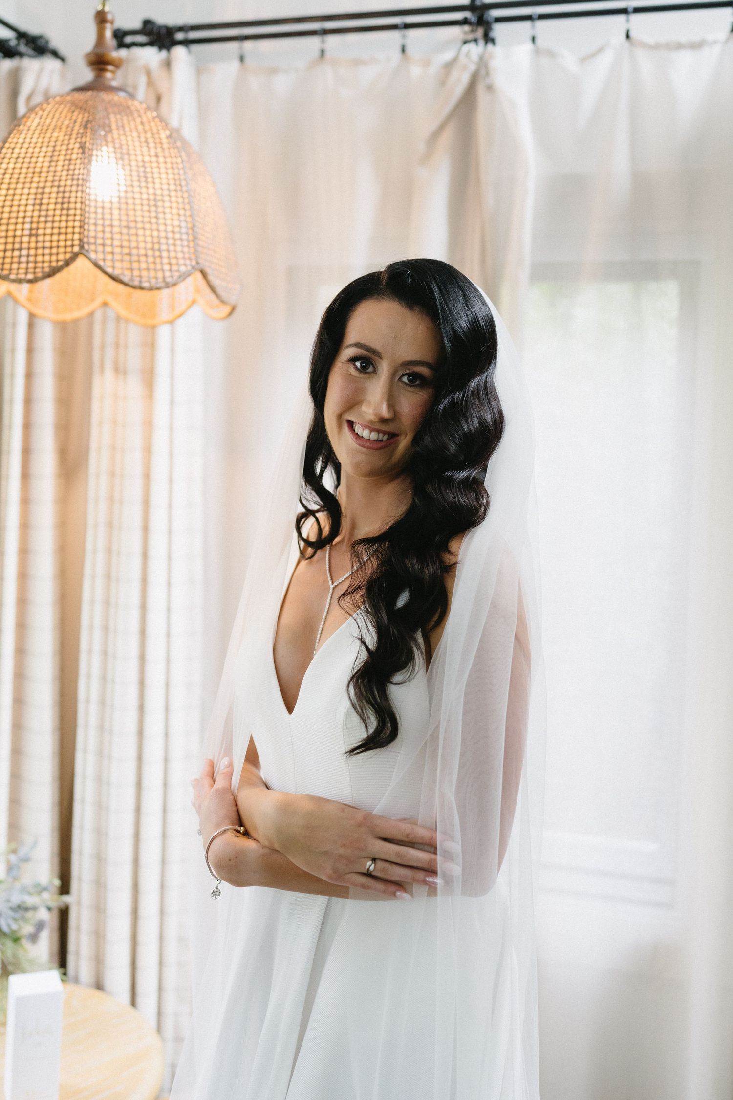 Bride in white v-neck gown poses by vintage lamp and sheer curtains.