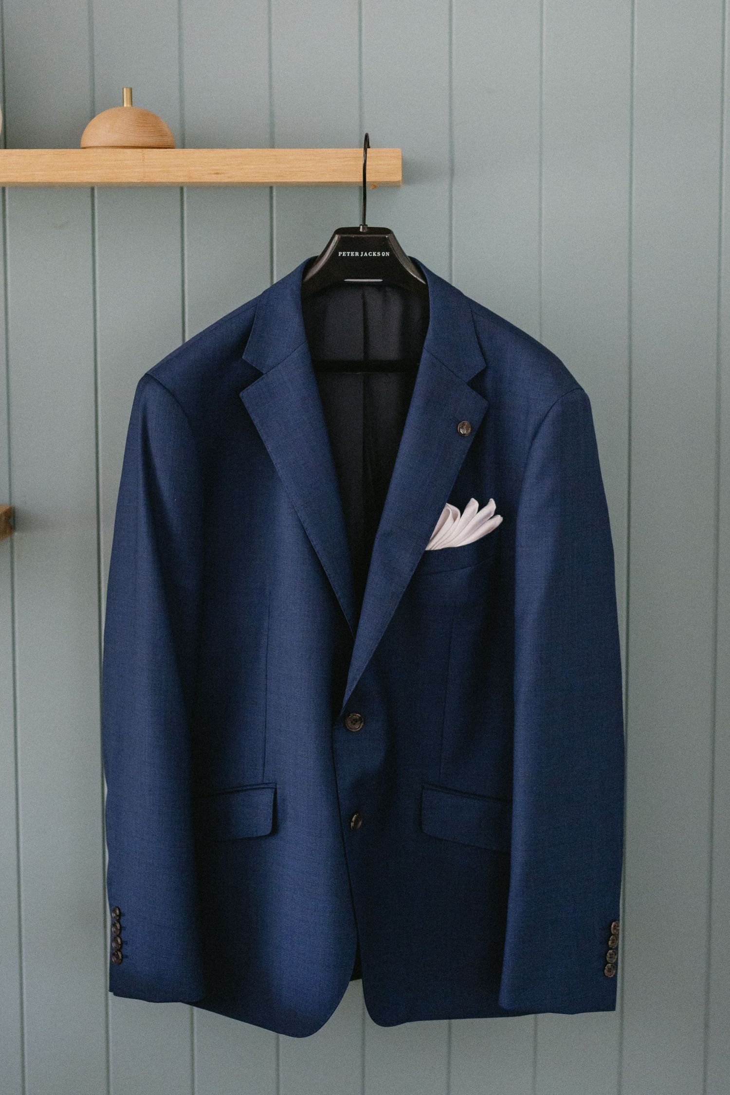 Navy blue formal suit jacket with white pocket square hanging on wooden hanger against gray wall.