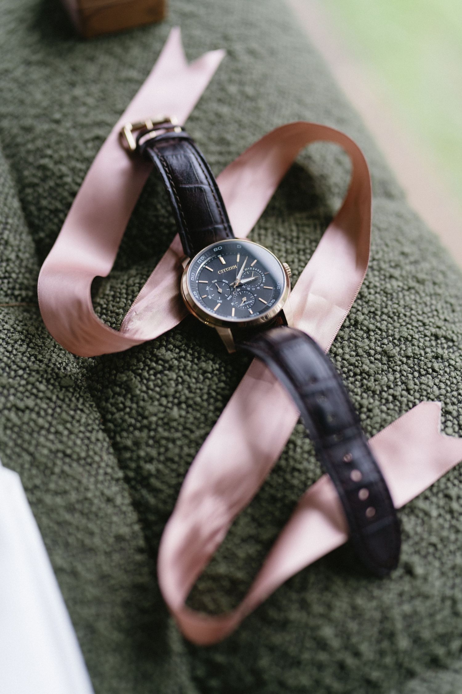 Elegant wristwatch with black leather strap and pink ribbon on green textile background.