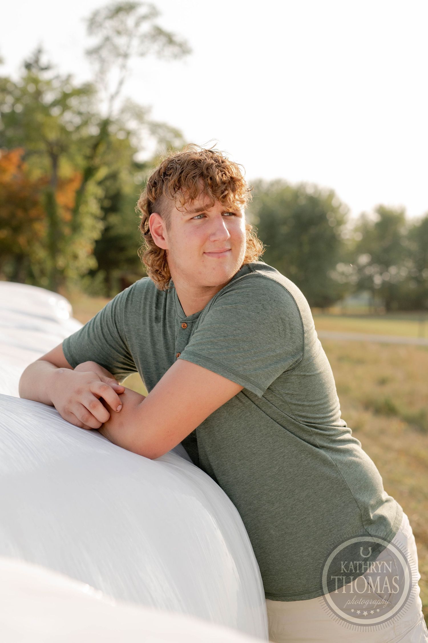 A person in a gray t-shirt leans against a white round bale of hay during golden hour.