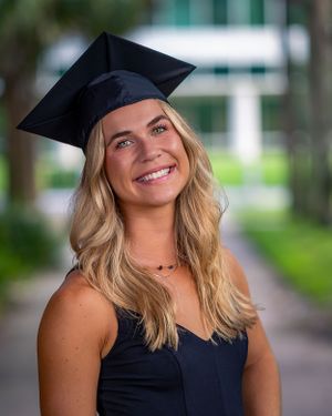 Graduate wearing a black cap and gown poses outdoors on a walkway with a bright smile and long blonde hair.