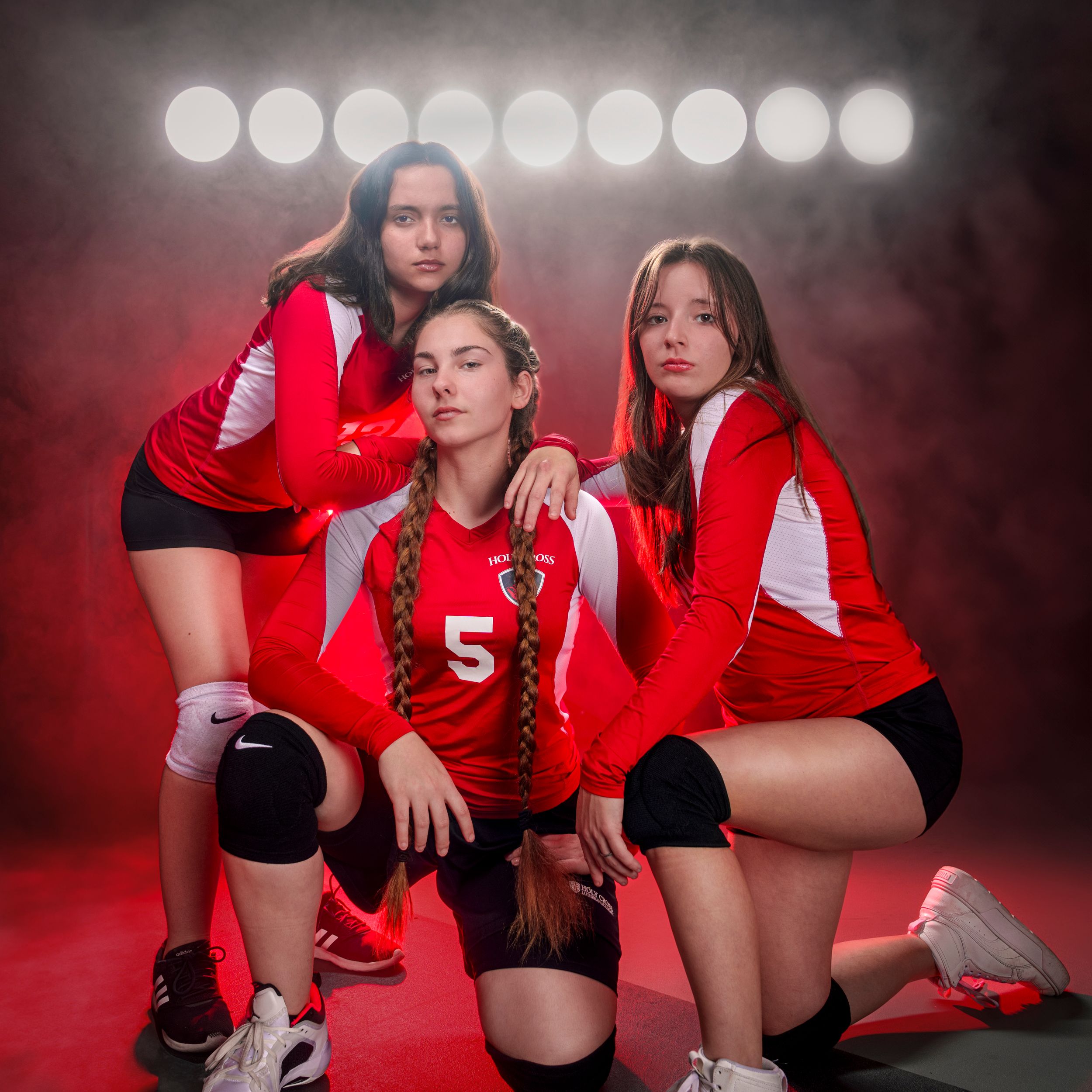 Three volleyball players in red and black uniforms pose dramatically with stage lighting and red backdrop.