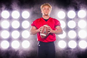 Football player in red jersey poses with ball against bright stadium lights in dramatic portrait.