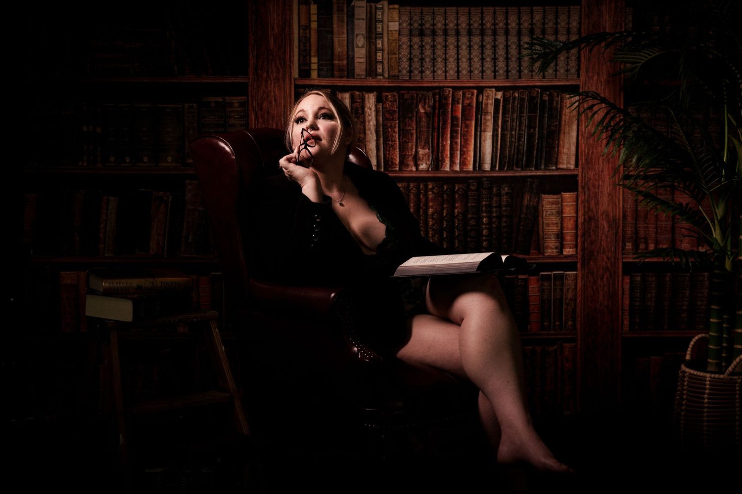Moody low-key portrait of a person in black dress lounging dramatically in vintage library setting.