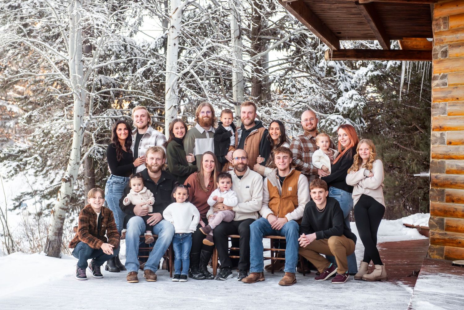 A large group poses together on a snowy winter porch of a log cabin surrounded by snow-covered trees.