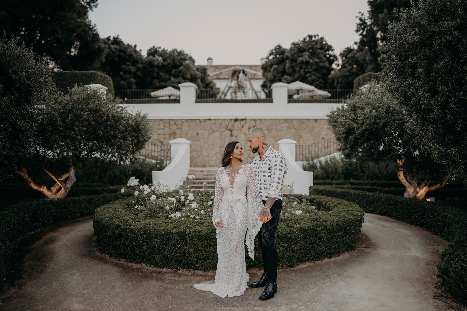 A romantic couple shares a tender moment in a beautifully landscaped garden with ornate hedges and architectural elements.