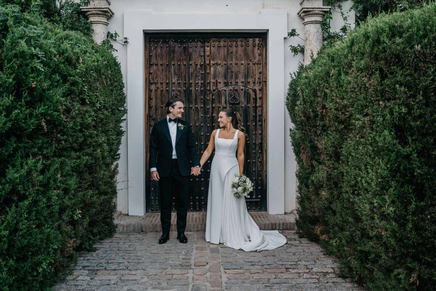 A couple in formal wedding attire stands between green hedges in front of an ornate wooden door at a historic venue.