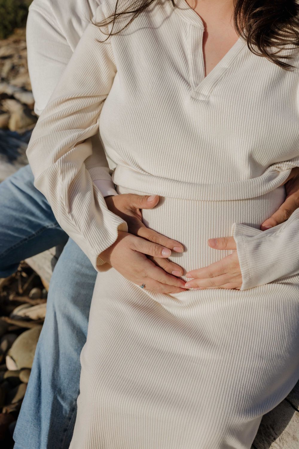 Close up of someone wearing a cream ribbed sweater dress and blue jeans with hands resting on their midsection.