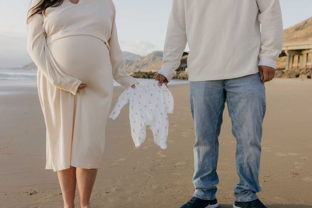 Expecting parents hold tiny white baby onesie while standing together on peaceful beach at sunset.