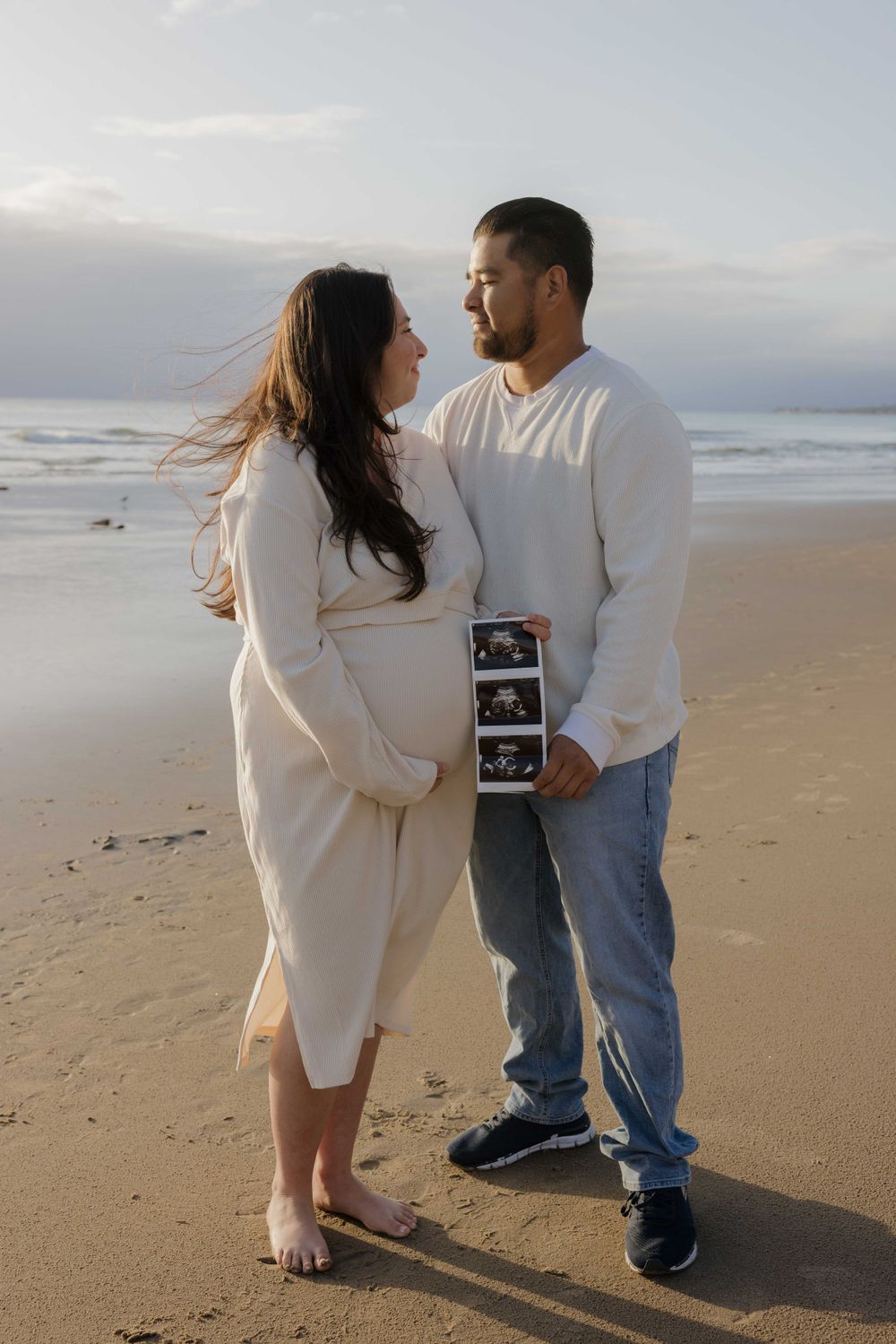 A couple shares an intimate moment on the beach while holding an ultrasound photo, announcing their pregnancy.