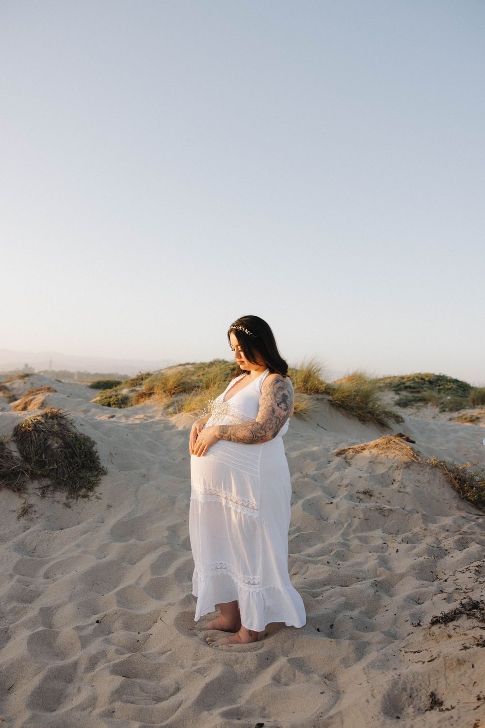 A pregnant woman in a flowing white dress stands serenely on sandy dunes at sunset against a pale sky.