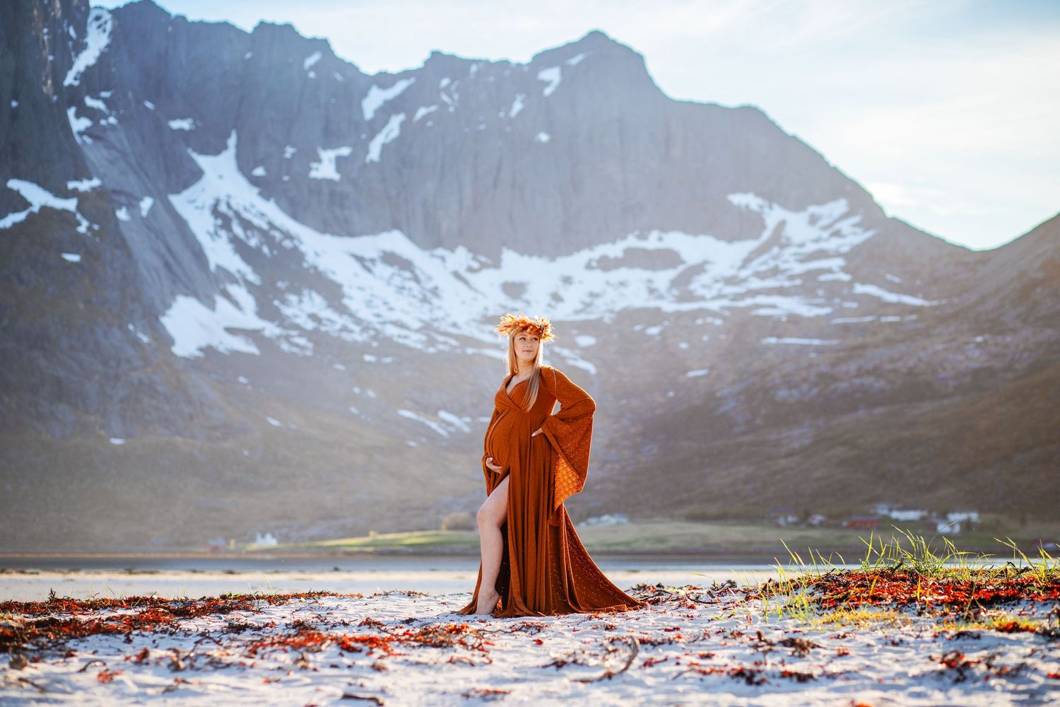 Woman in rust orange gown with floral crown poses on snowy beach near mountains.