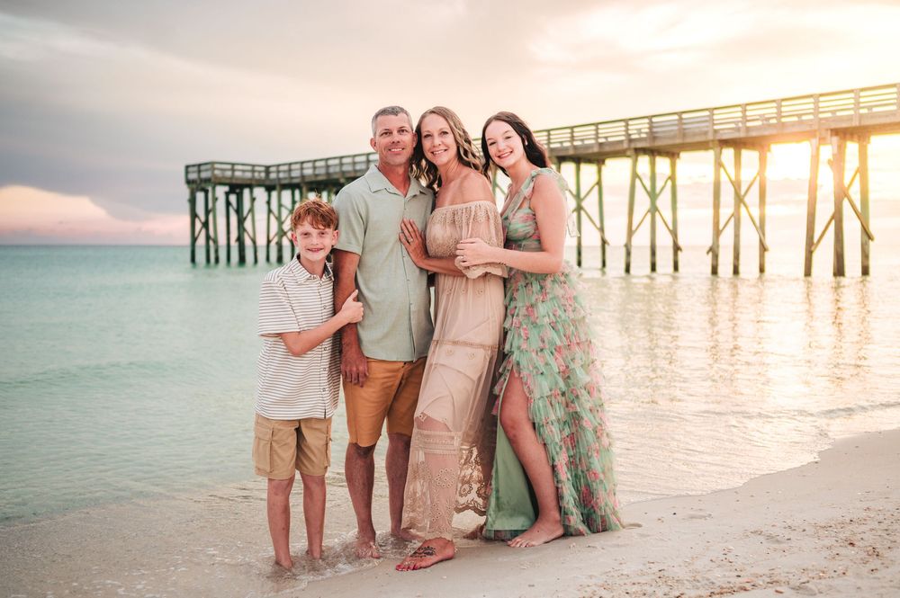 A group of people standing together on a beach at sunset with a wooden pier in the background.