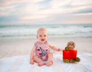 Adorable baby wearing blue plaid outfit sits for multiple beach portraits next to stylish hat and red plush toy at sunset.
