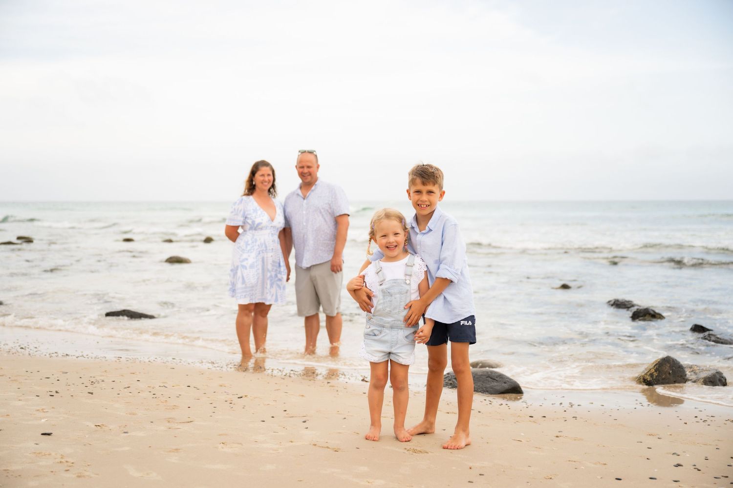 A family in white clothing stands together on a beach with ocean waves and rocks in the background.