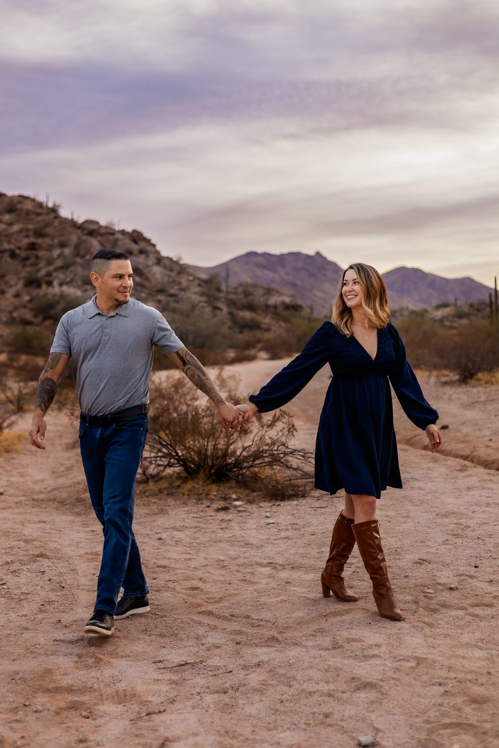 Couple holding hands while walking through a desert trail with mountains in the background at sunset in Phoenix.