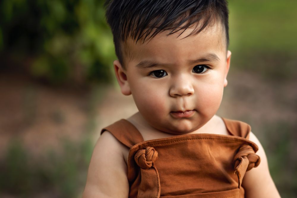 Baby in brown overalls looking at camera with soft natural lighting for his first birthday milestone photos.