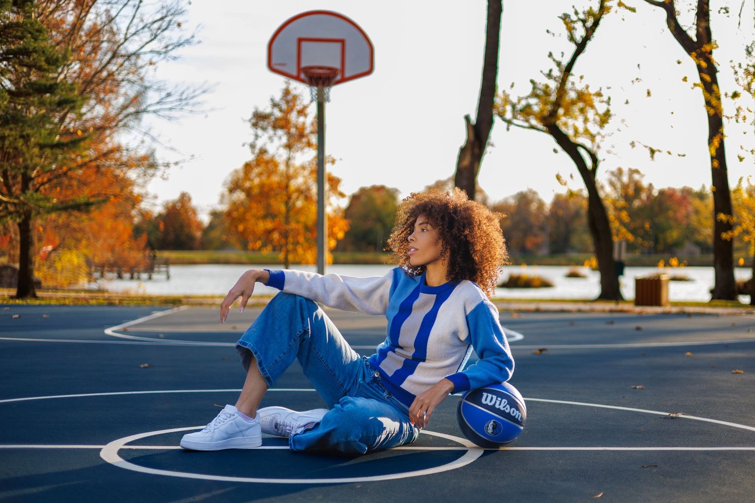 Basketball player with curly hair relaxes on outdoor court during autumn sunset in blue and white uniform.