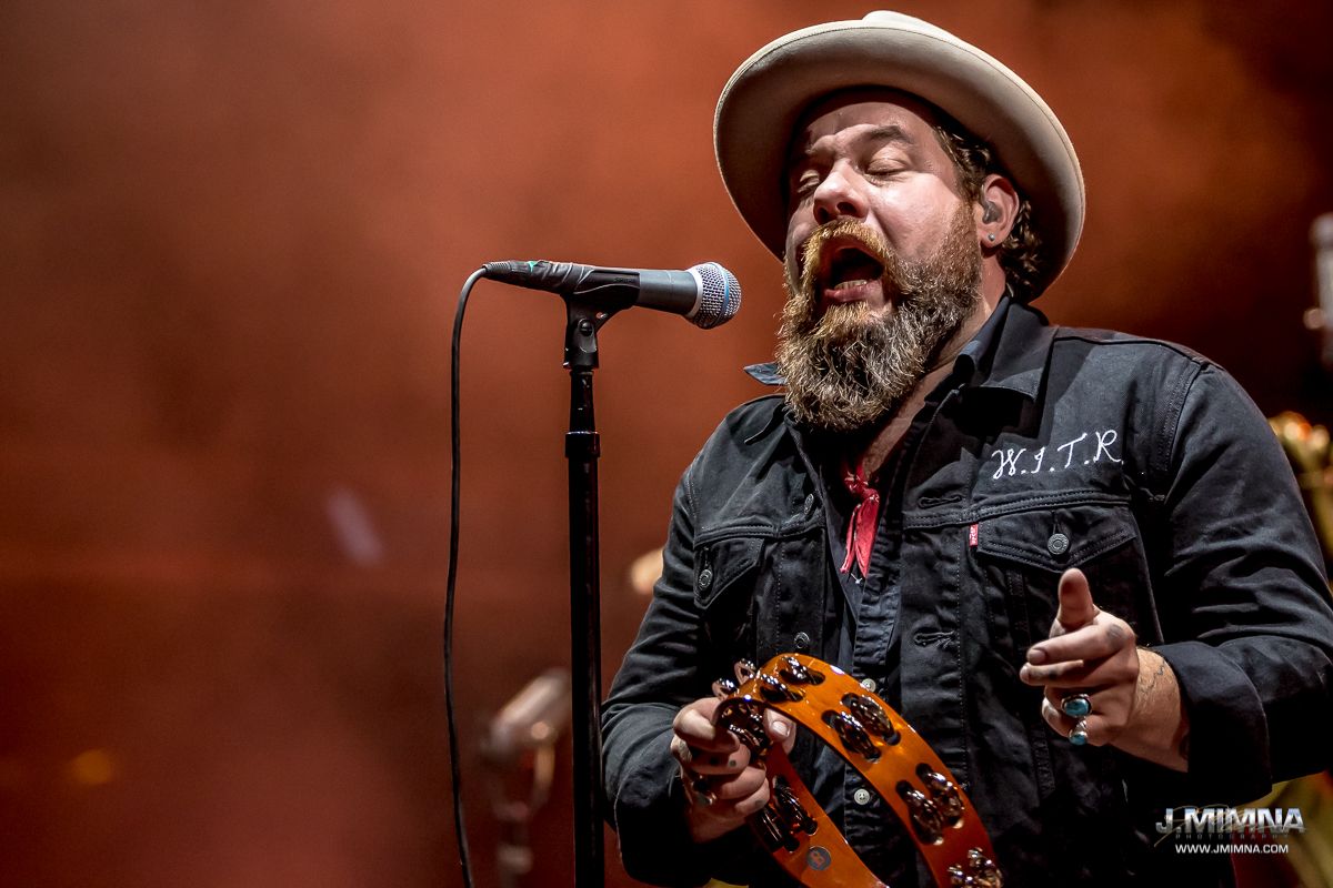 A passionate singer in a black jacket and cowboy hat performs energetically with a microphone on stage with orange lighting.