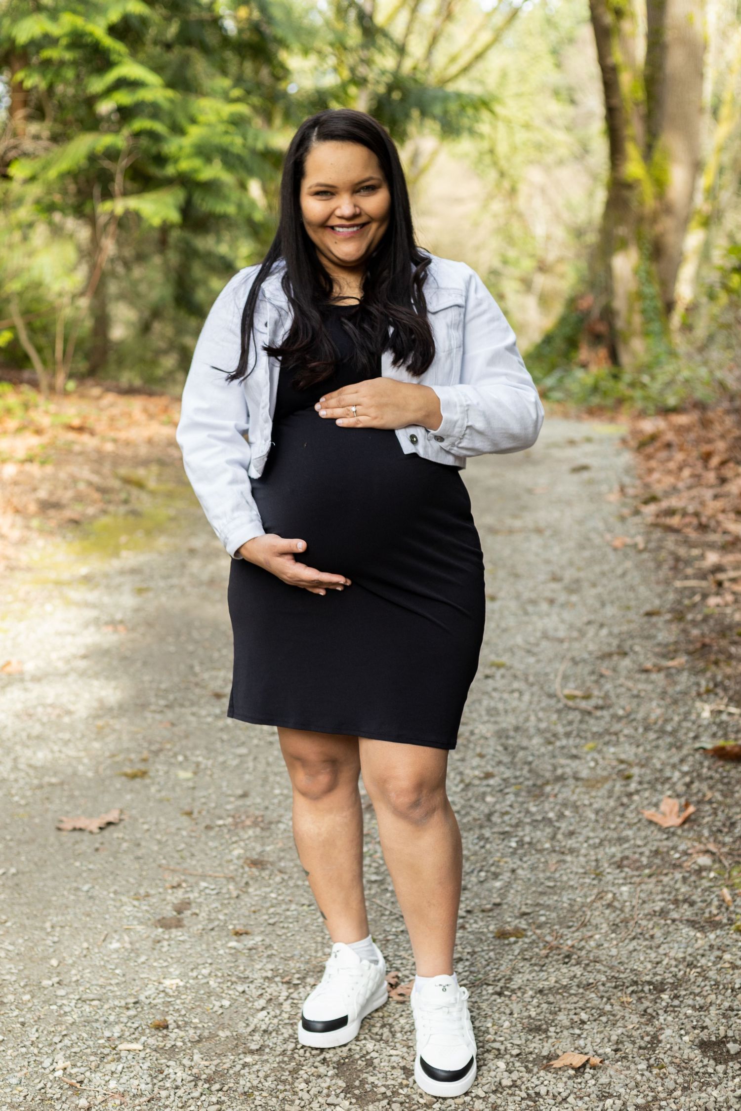 Expectant mother in black dress and white jacket smiles on forest path.