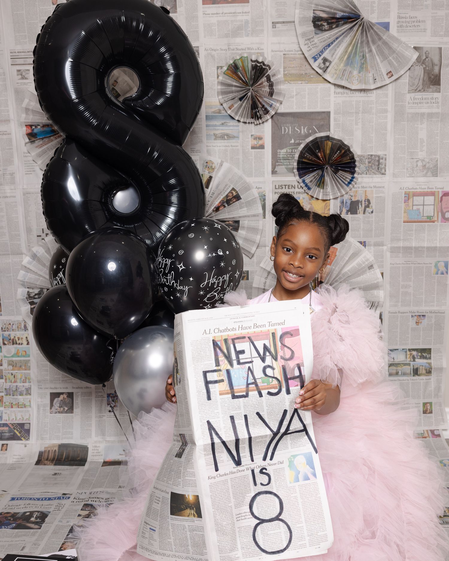 A festive birthday setup with large black number 8 balloon, silver accents, and newspaper backdrop with a handmade sign.