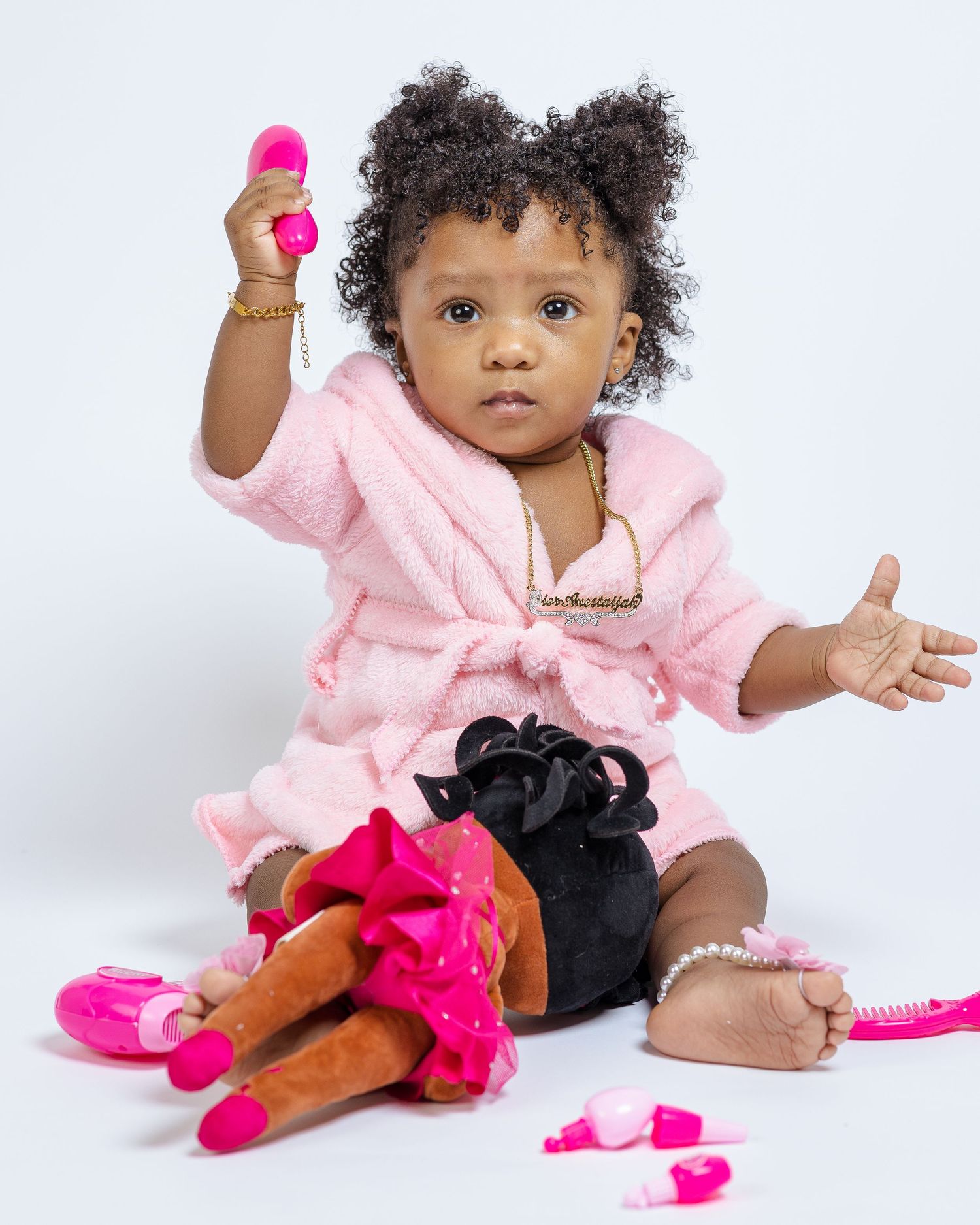 Small child in pink bathrobe plays with colourful toys and accessories on white background.