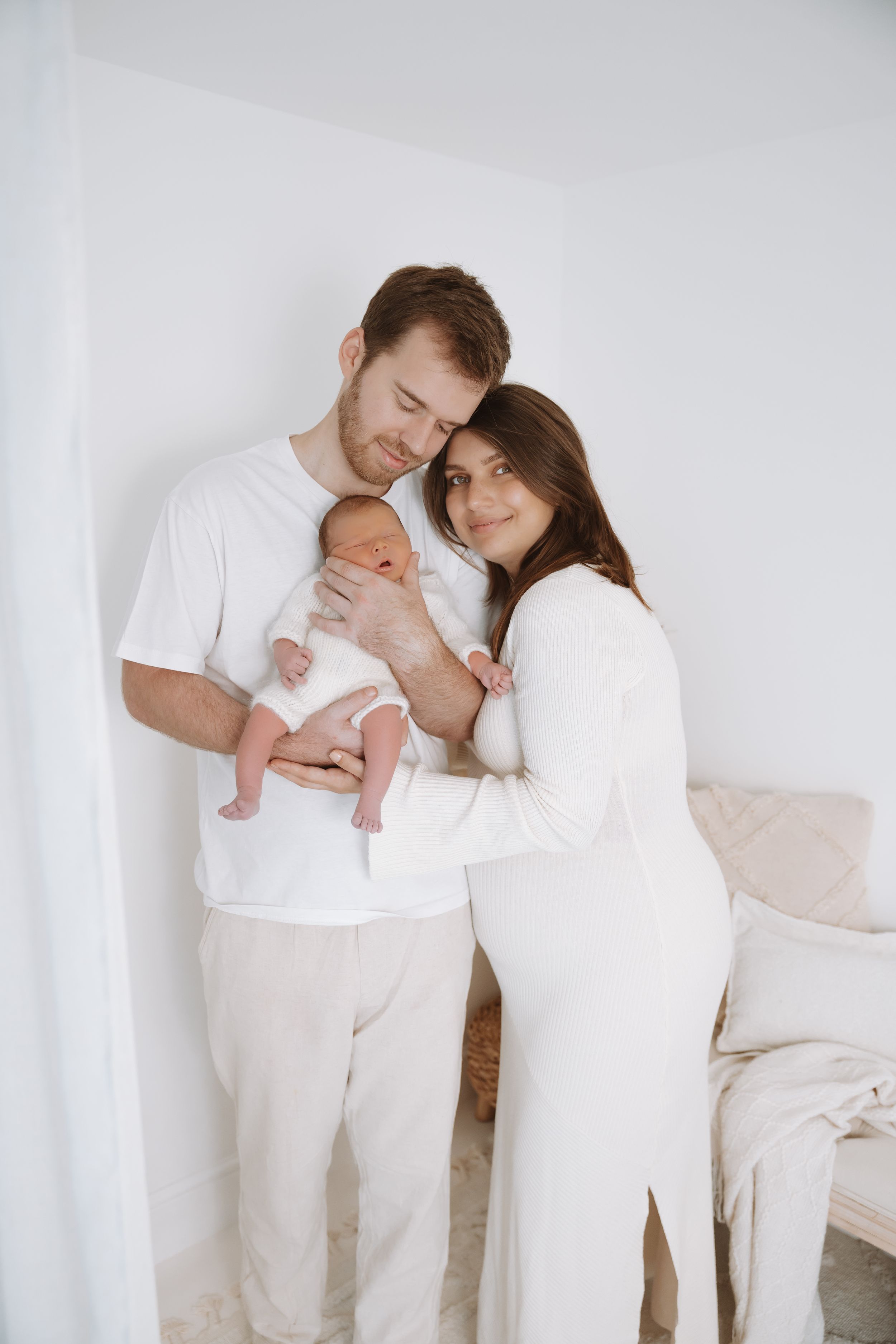 Family wearing all white outfits embraces tenderly in minimalist studio setting.
