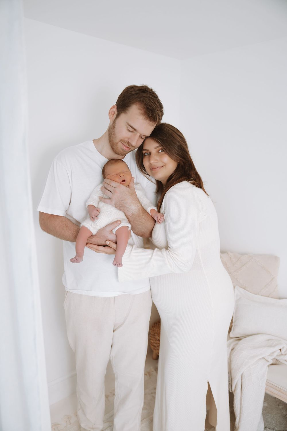 Family wearing all white outfits embraces tenderly in minimalist studio setting.