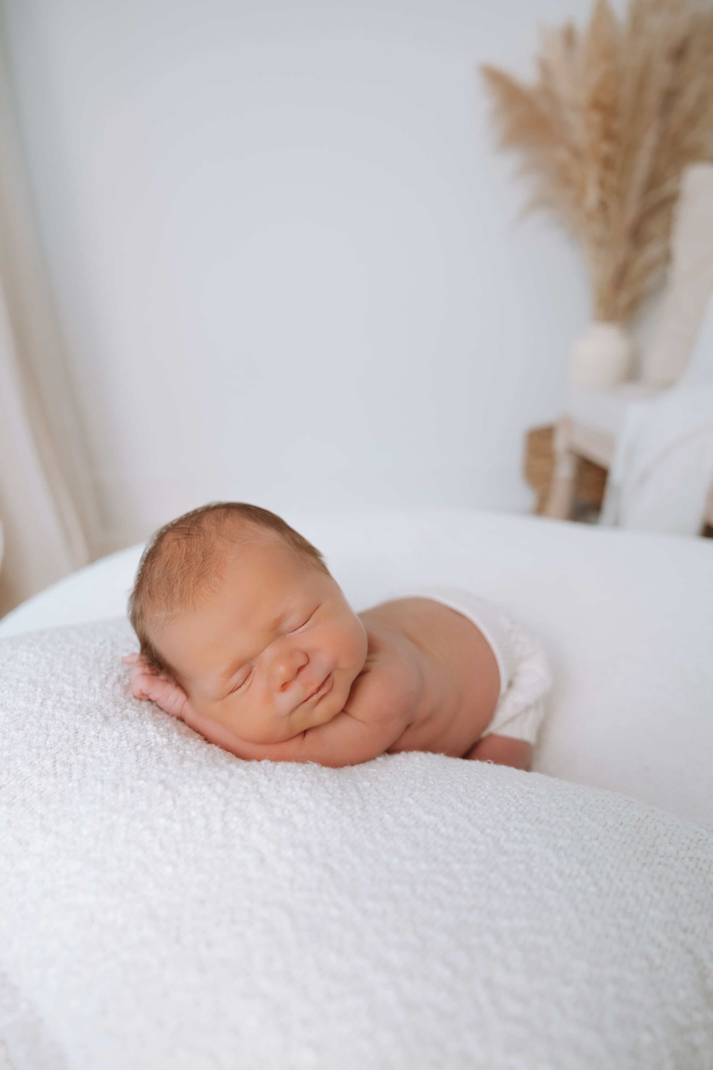 A peaceful sleeping newborn rests on soft white textured bedding with dried pampas grass decor in the background.