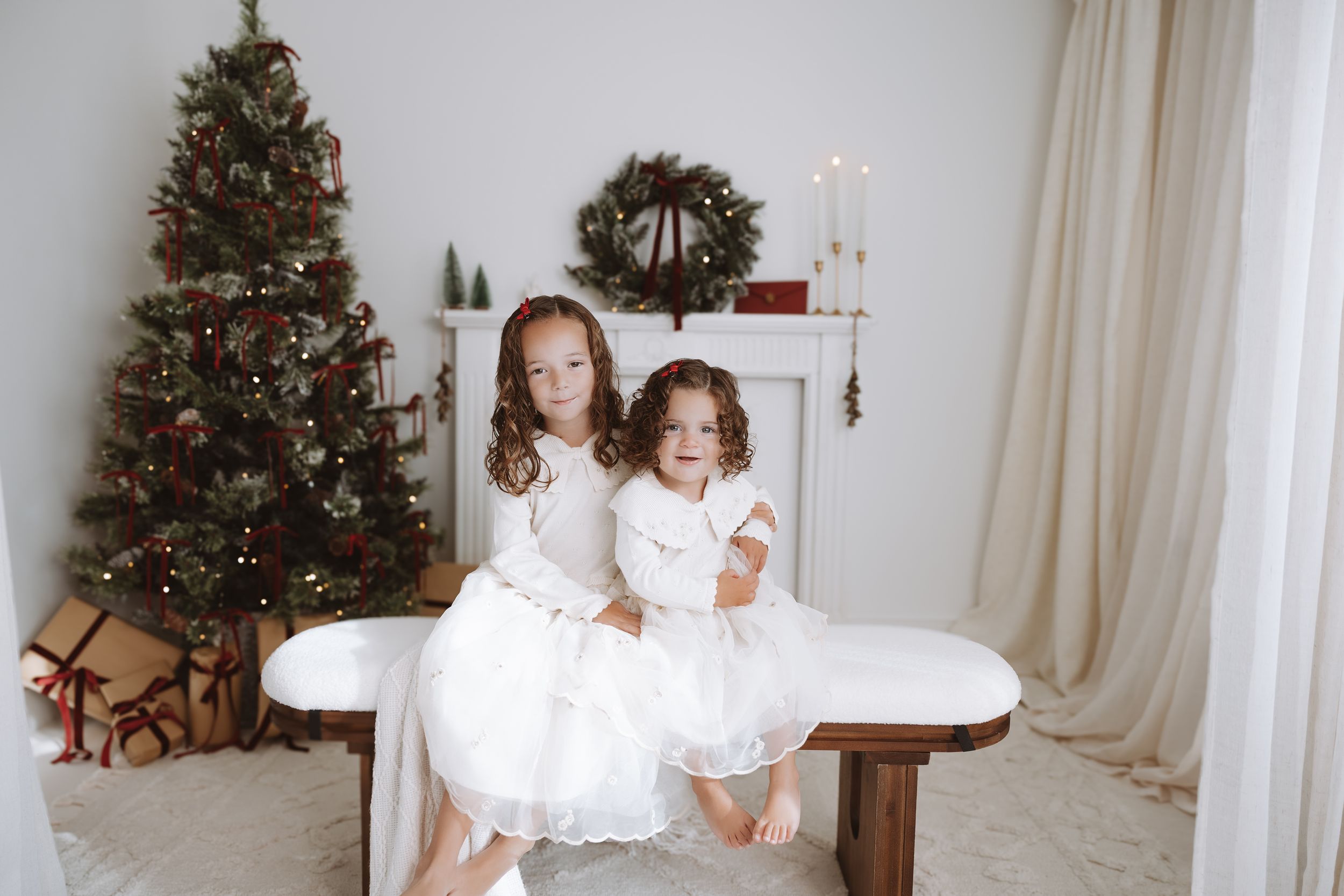 Two young girls in white dresses sit together on a bench near a decorated Christmas tree and fireplace.