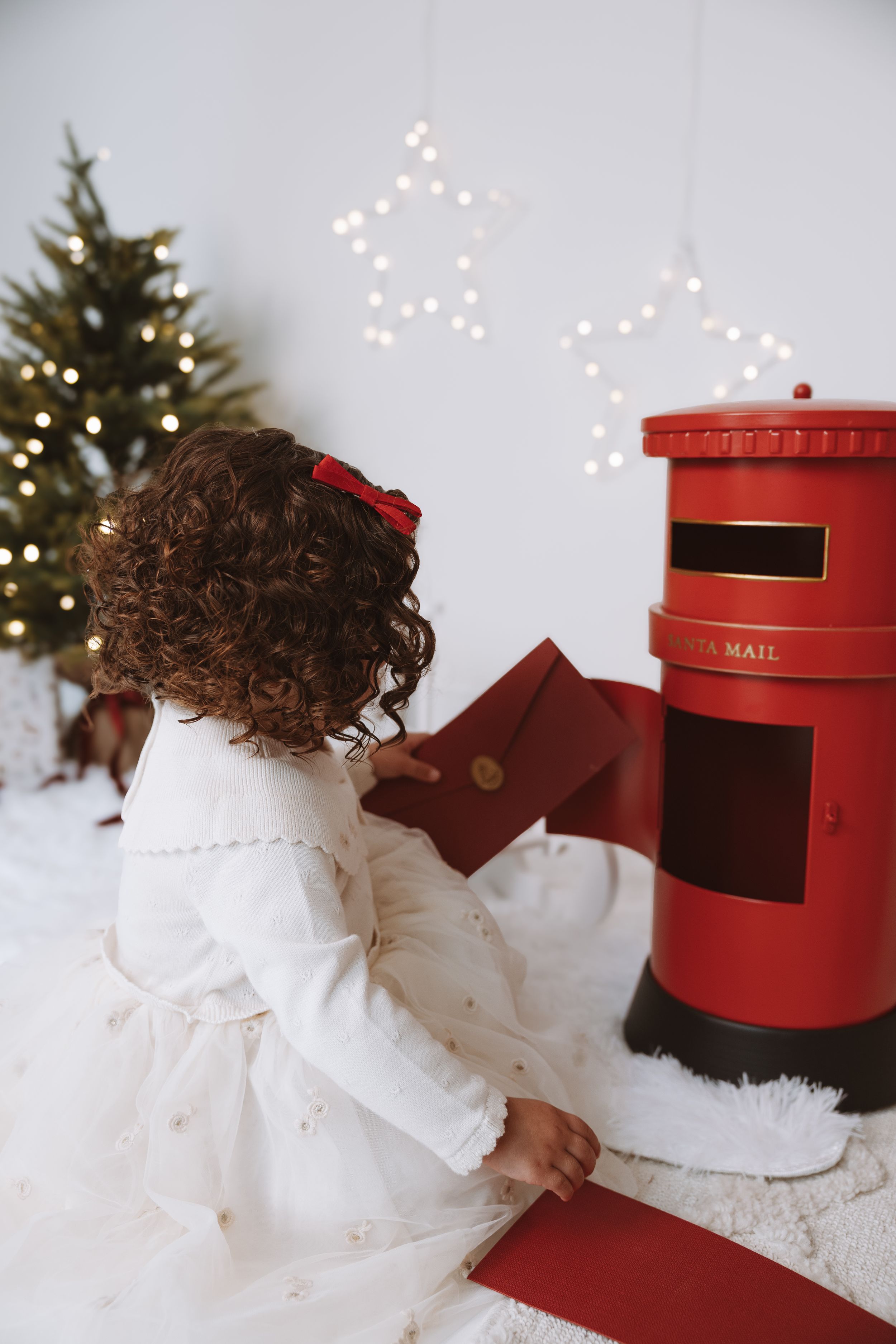 Young girl in white dress posting a red envelope into a Santa Mail box beside a decorated Christmas tree.