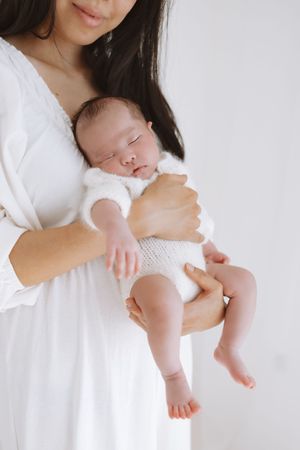 A sleeping newborn baby in white clothing is cradled gently against a white dress.