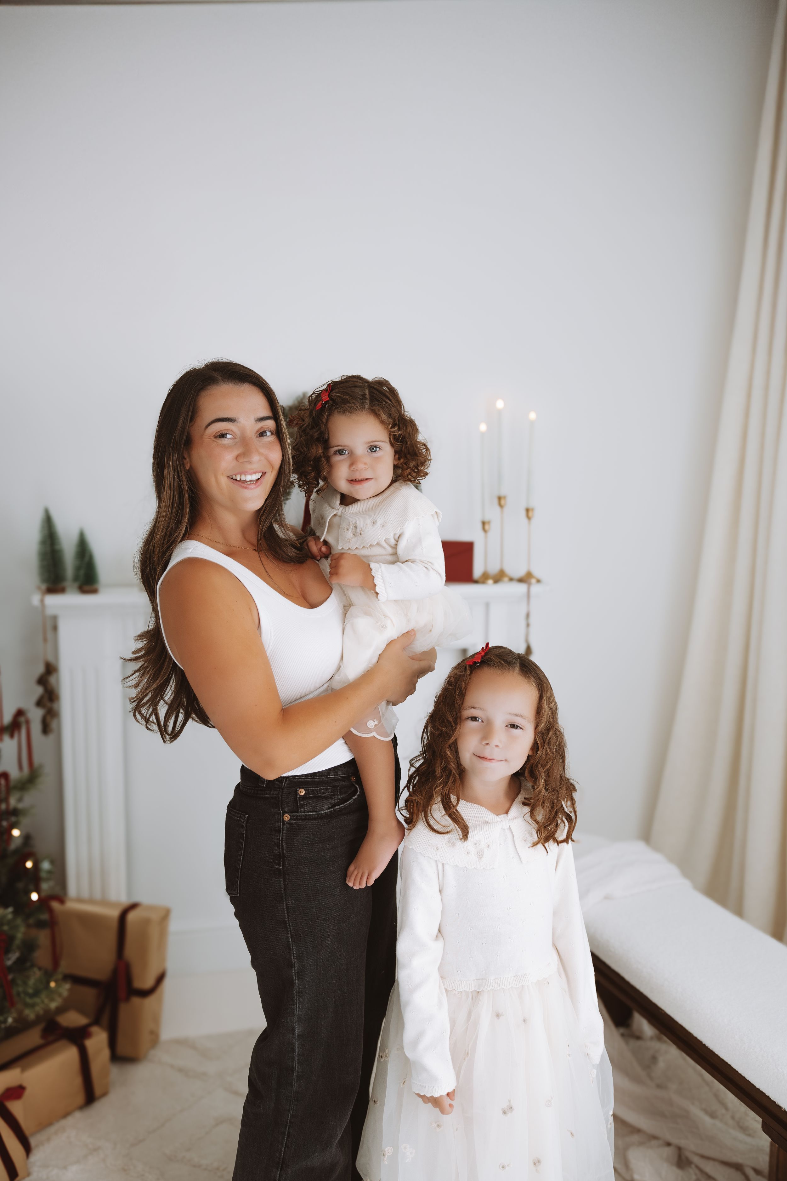 A smiling woman holds a toddler while a young girl in a white dress stands beside her in a Christmas decorated room.
