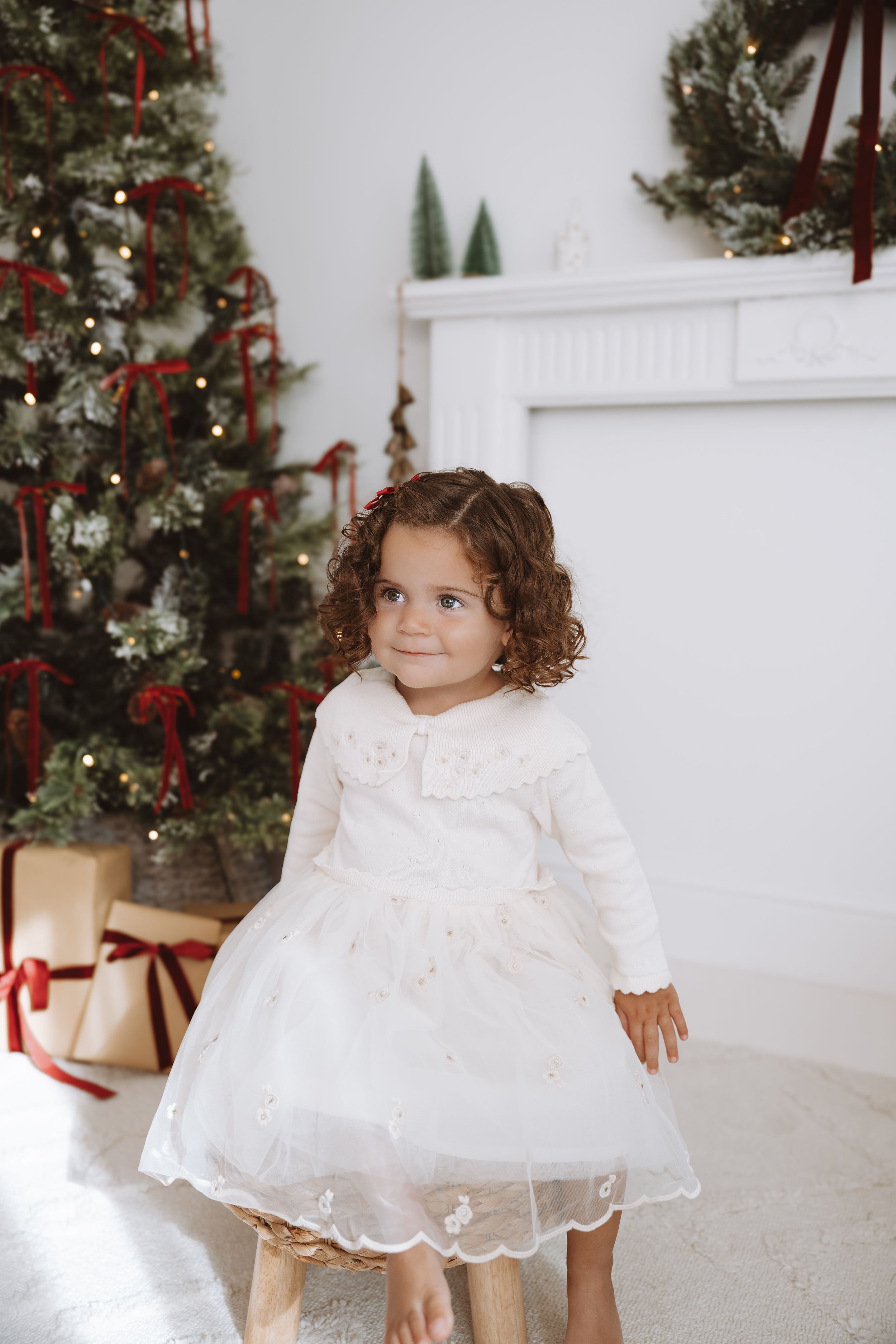 Adorable toddler girl in white dress poses near decorated Christmas tree with gifts and festive wreath.