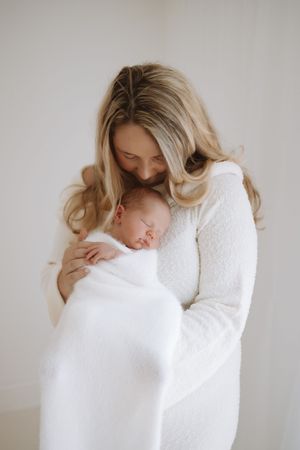 Mother in white sweater gently embraces and kisses sleeping newborn wrapped in white blanket.