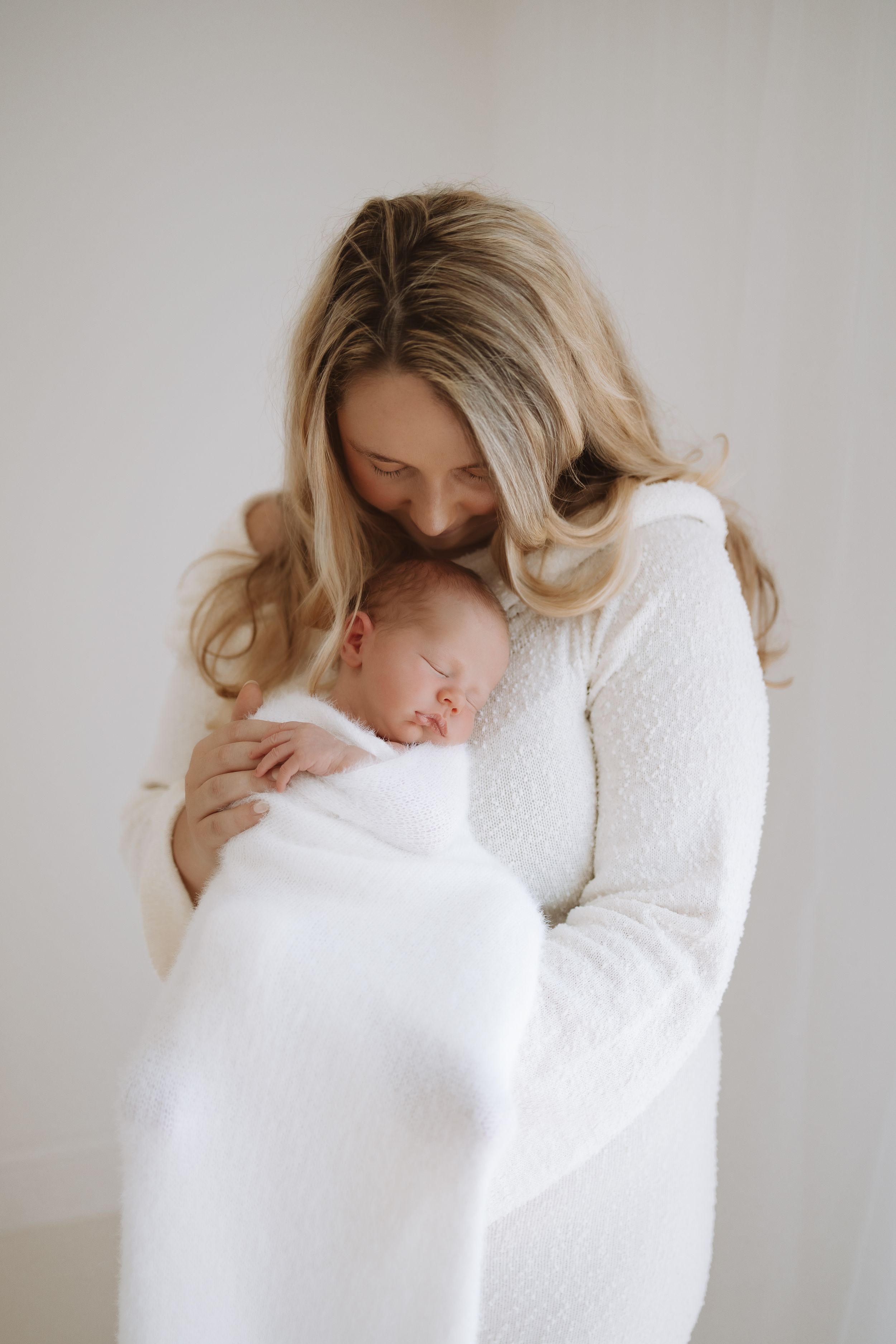 Mother in white sweater gently embraces and kisses sleeping newborn wrapped in white blanket.