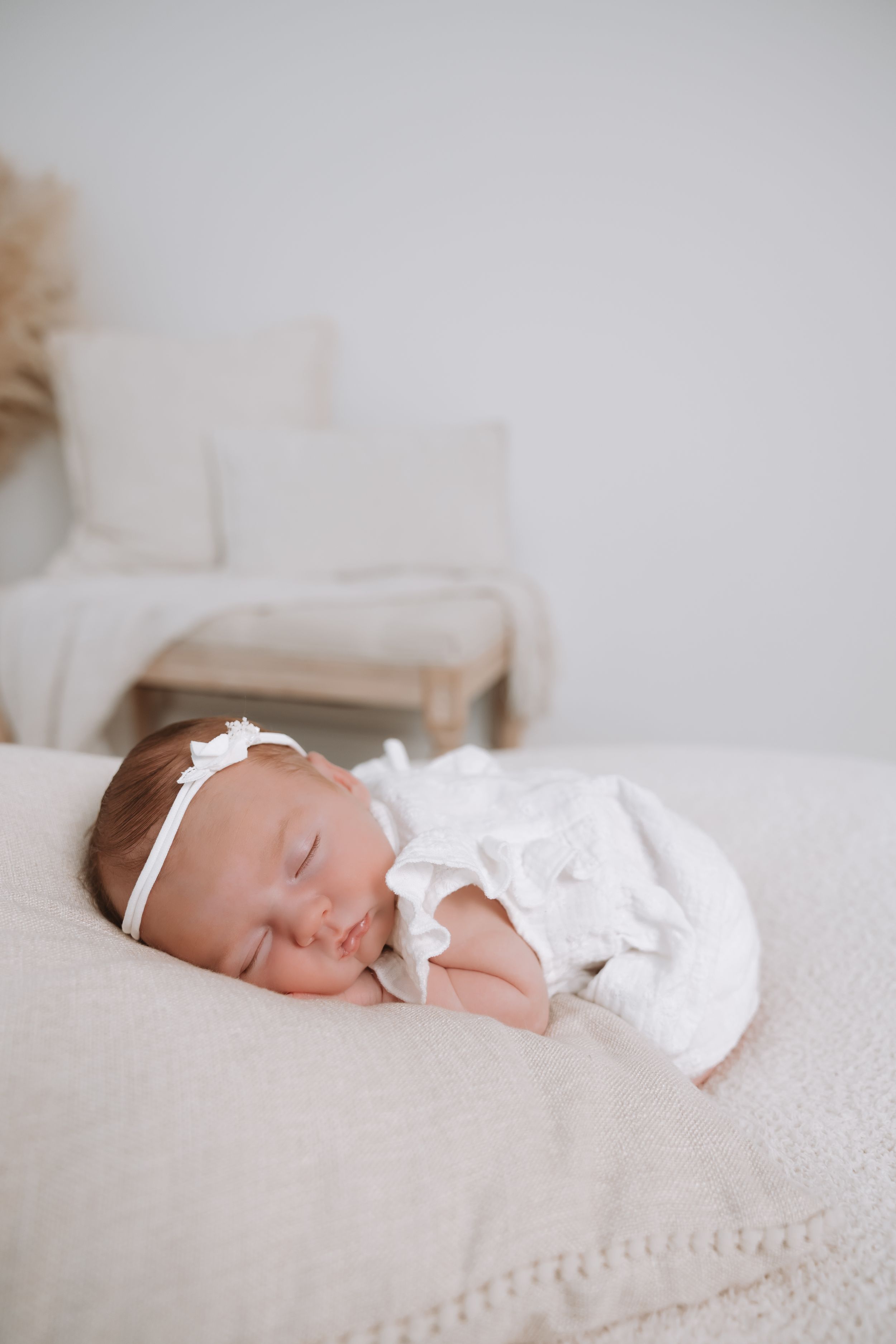 Peaceful newborn baby girl sleeping in white lace outfit and headband on a soft cream blanket.