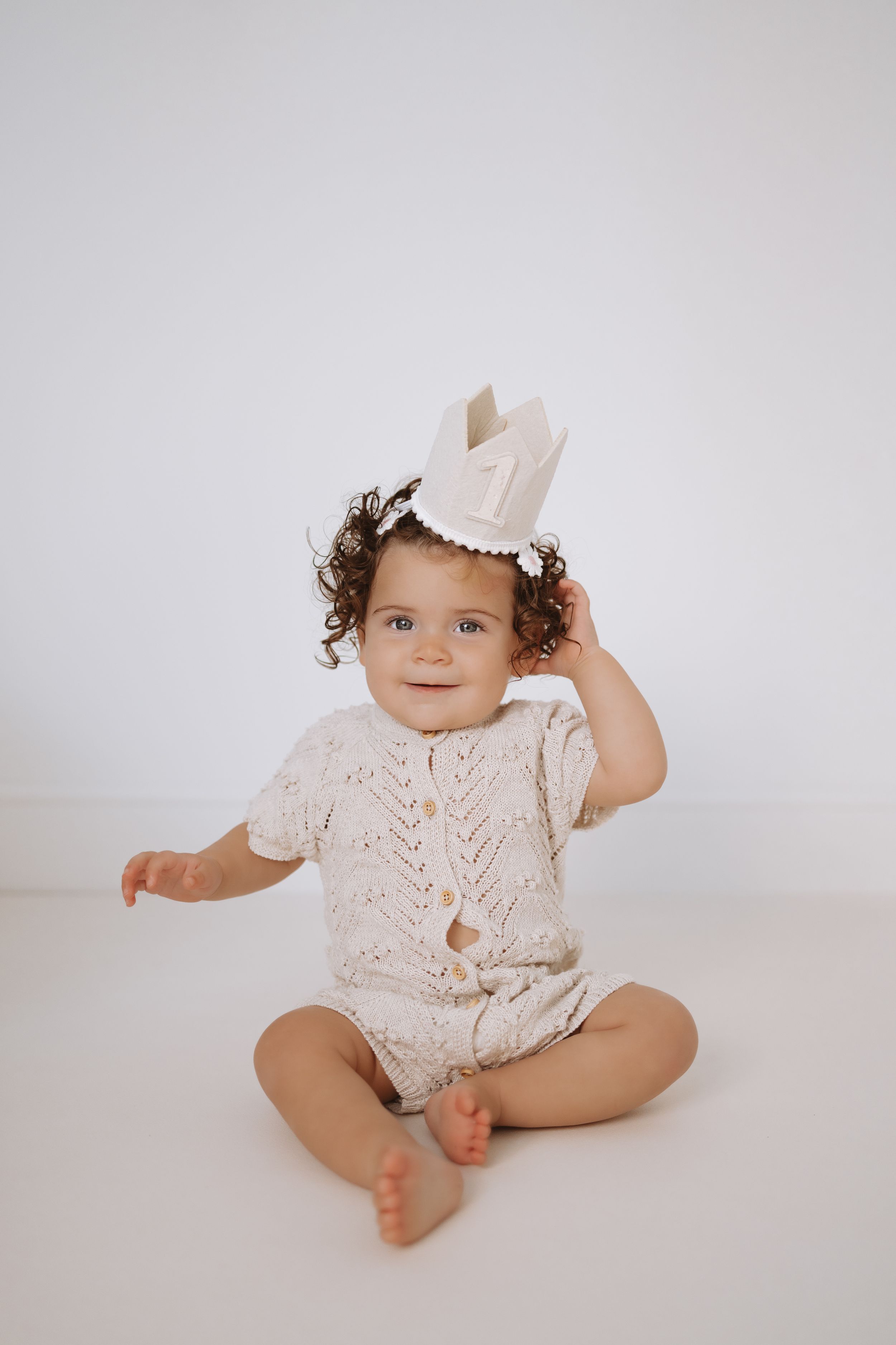 A baby wearing a white lace romper poses on a light background in a series of sweet studio portraits.