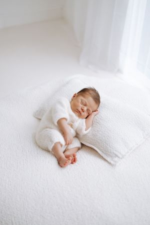 A sleeping baby in white clothing rests peacefully on crisp white bedding in a gentle, minimalist setting.