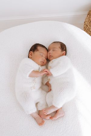 Sleeping newborn twins in white outfits lying close together on soft white bedding.