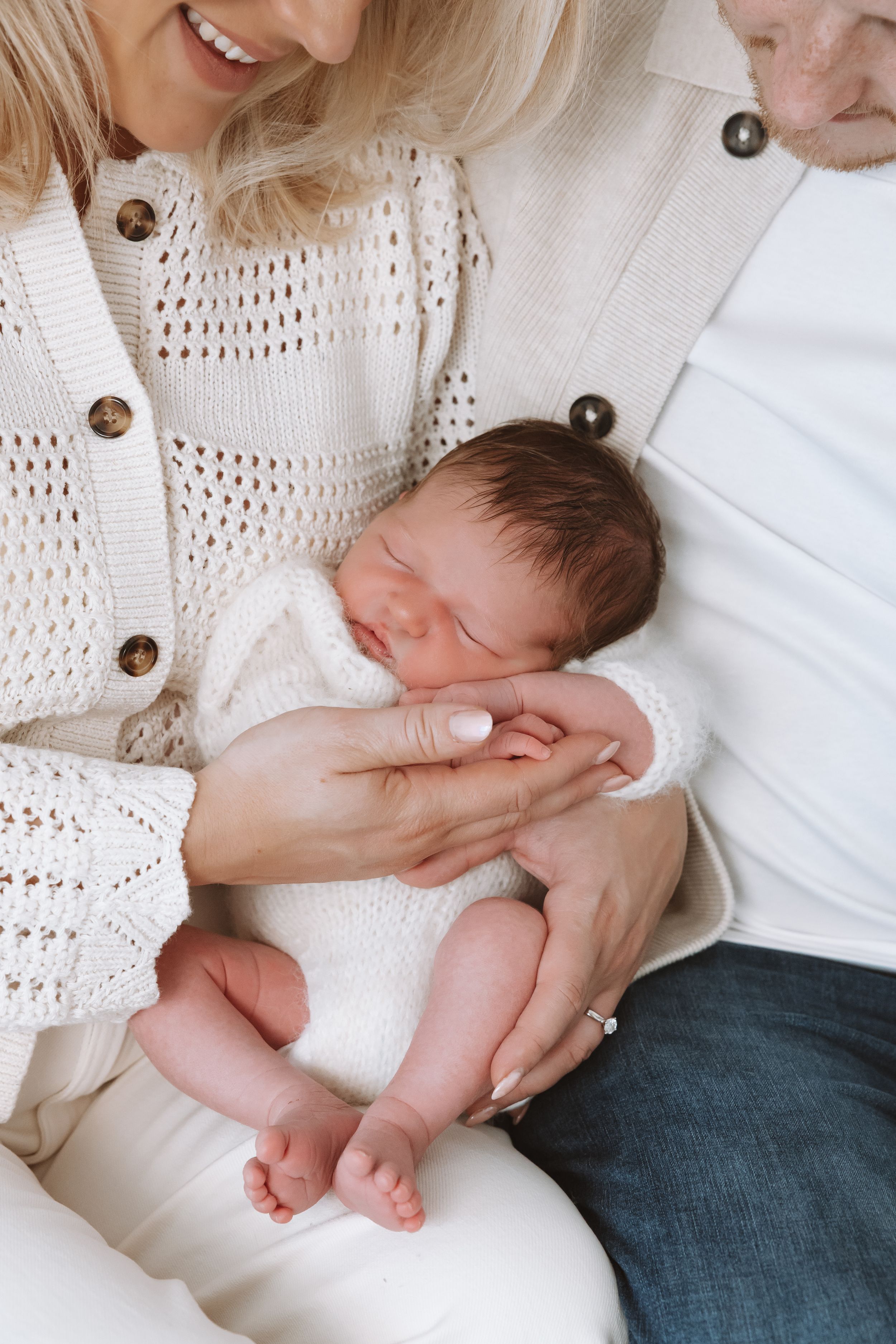 A sleeping newborn baby rests peacefully in a caregiver's arms while wearing white against a cream knit sweater.