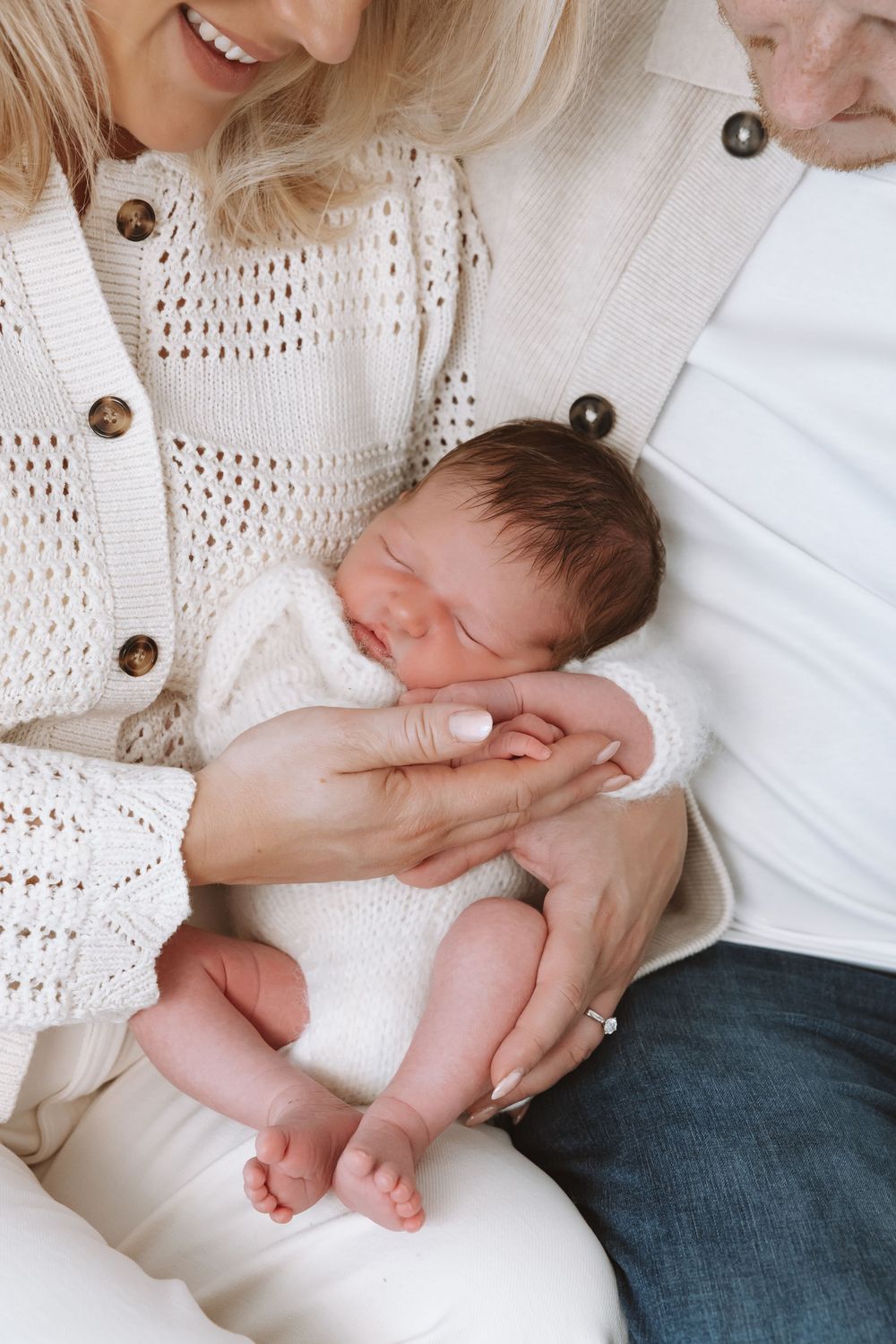 A sleeping newborn baby rests peacefully in a caregiver's arms while wearing white against a cream knit sweater.