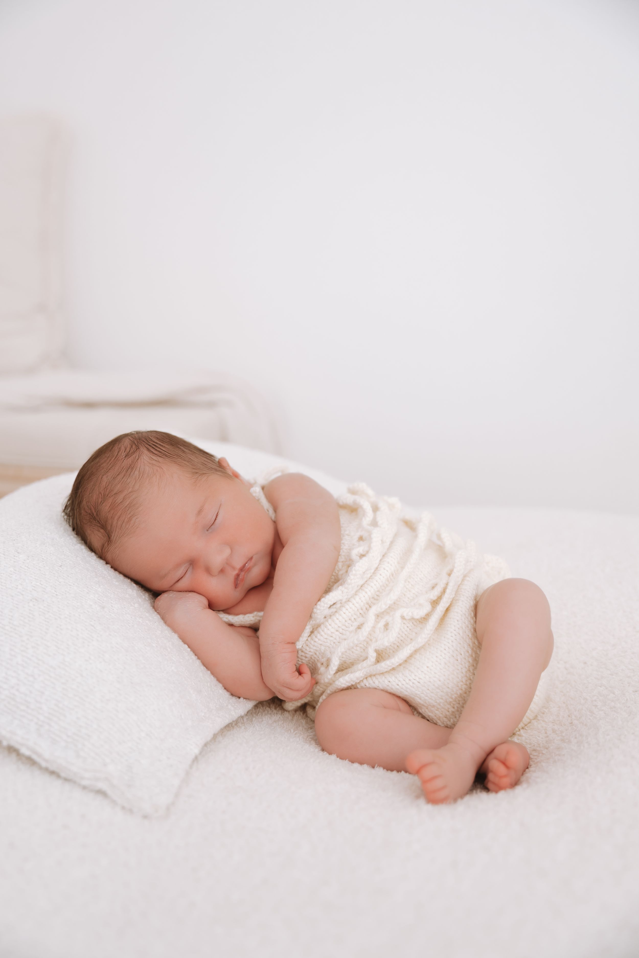 Peaceful newborn baby sleeping on a white pillow wearing a cream knit romper in a bright, minimalist setting.