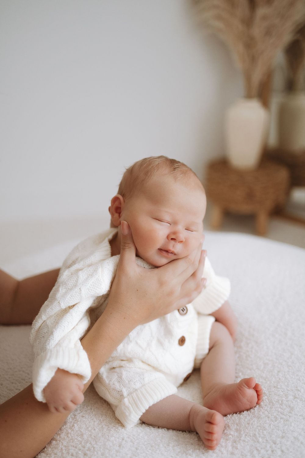 Sleeping newborn baby wearing white lace outfit lying on textured white blanket in natural light setting.