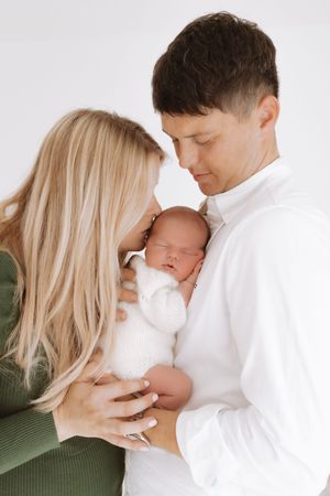 A tender family moment as parents embrace their newborn baby in white against a light background.