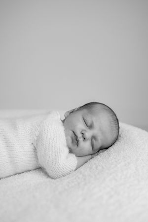Peaceful sleeping newborn baby wrapped in soft white knitted blanket against light background.