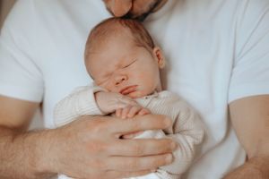 Peaceful newborn sleeping in white onesie while being held against white shirt.