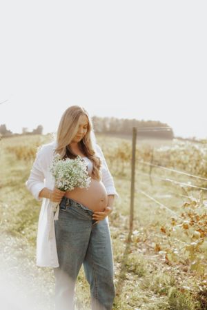 Expectant mother in white dress holding baby's breath flowers in sunny field setting.